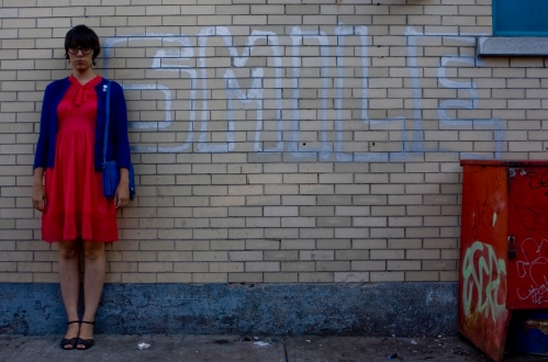 a photo of julia, a young femme presenting person, standing next to a silver graffiti that reads SMILE. she is not smiling.