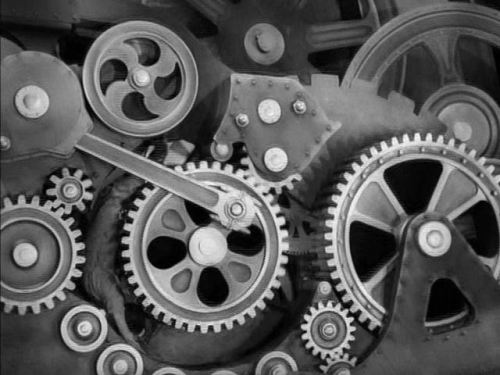 A film still from Charlie Chaplin's Modern Times (1936) showing Chaplin's character stuck in the gears of a giant machine